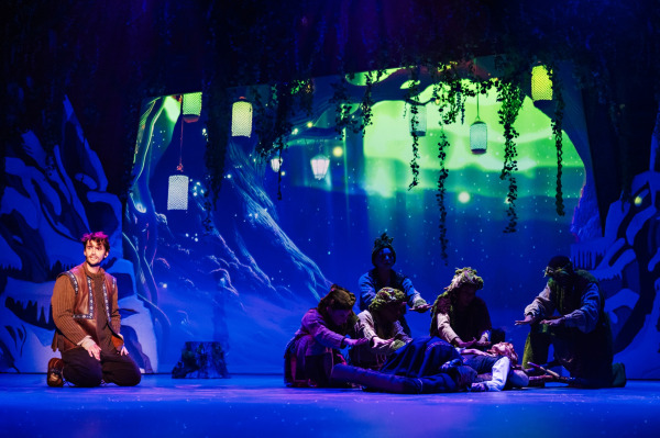 A performer in a sparkling ice-blue gown sings before a towering frozen palace projection featuring a large crystalline snowflake. At left, an enlarged translucent image of the performer appears against a dark, starry background.