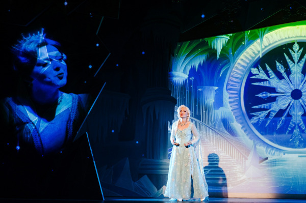 A performer in a sparkling ice-blue gown sings in front of a towering frozen palace projection dominated by a large crystalline snowflake. At left, an enlarged translucent image of the same performer appears against a dark, starry background, creating a dramatic layered visual effect.