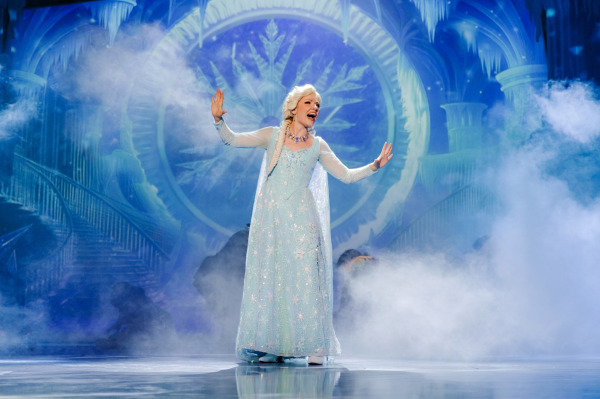 A performer in a sparkling ice-blue gown sings centre stage with her arms raised as mist swirls around her. An elaborate frozen palace glows behind her in shades of blue, with icy columns, sweeping staircases, icicles, and a large crystalline snowflake.
