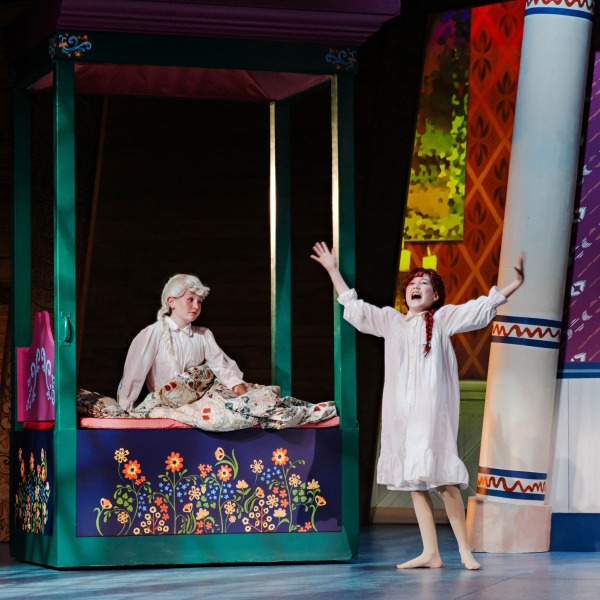 Two young performers in white nightgowns appear in a colourful bedroom scene. One sits beneath a patterned quilt on a tall green bed decorated with painted flowers, while the other stands barefoot beside the bed with both arms raised.