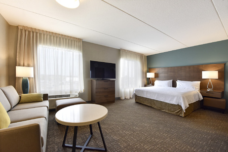 Spacious hotel studio suite featuring a king-sized bed with white linens, two bedside tables with modern lamps, a flat-screen TV on a wooden dresser, a beige sofa with green accent pillows, a round coffee table, and large windows with sheer curtains